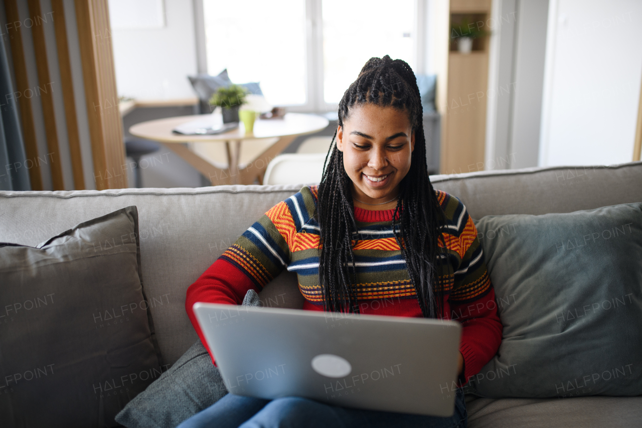 A happy teenage girl student on sofa indoors at home, using laptop.