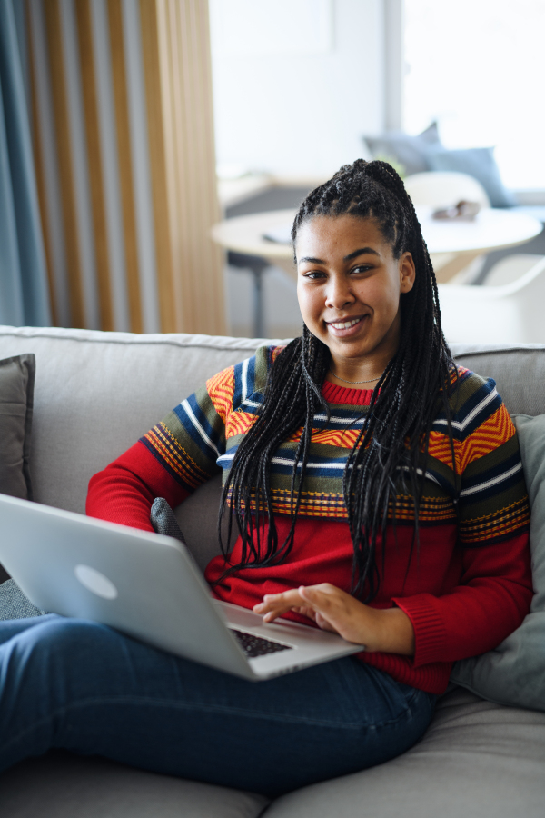 A happy teenage girl student with laptop on sofa indoors at home, looking at camera.