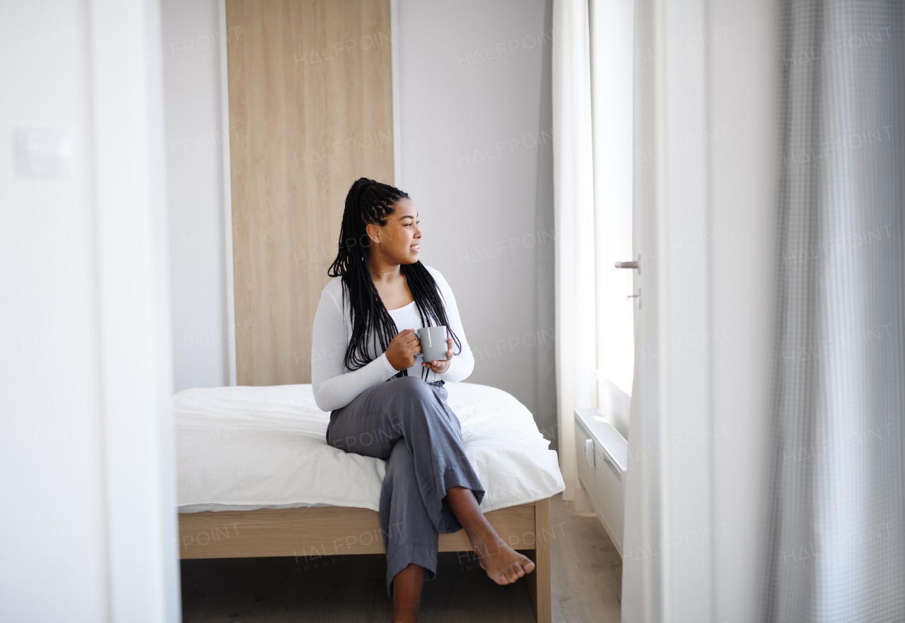 Portrait of happy teenage girl with cup of tea sitting indoors in bedroom at home, looking out of window.