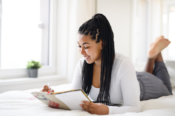 A happy teenage girl student lying indoors on bed at home, reading book and making notes.