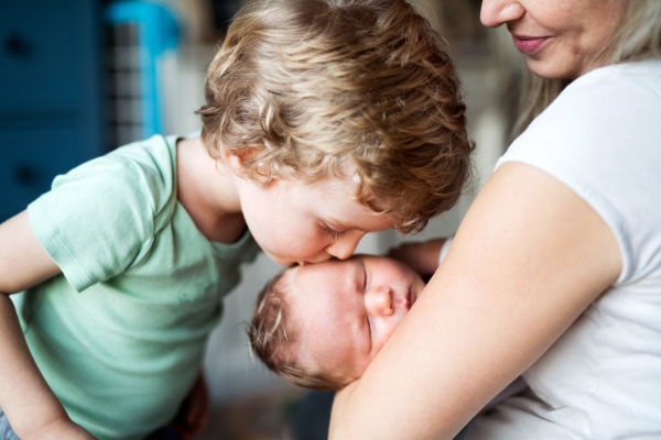 A small boy kissing a sleeping newborn baby brother at home, a mother holding him.