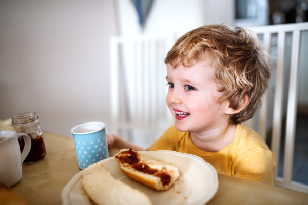 A front view of a happy toddler boy sitting at the table at home, eating.