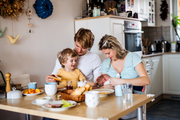 Young parents with a newborn baby and small toddler son sitting at the table at home.