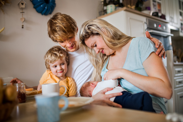 Young parents with a newborn baby and small toddler son sitting at the table at home.