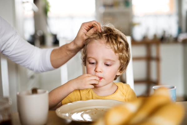 A front view of a sad toddler boy sitting at the table at home, eating.