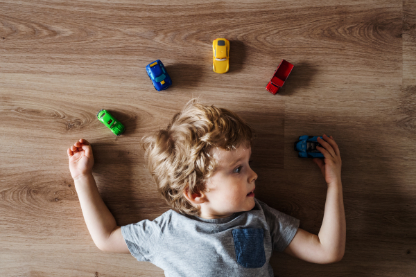 A top view of a toddler boy with toy cars lying on the floor at home.