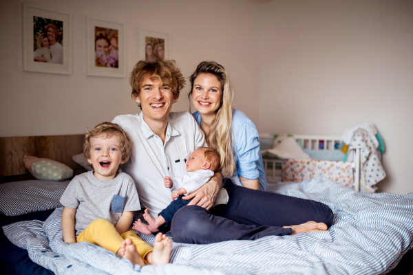 Portrait of young parents with a newborn baby and small toddler son sitting on bed at home.