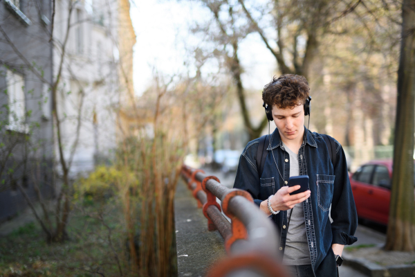 A portrait of young man commuter with headphones walking outdoors in city, using smartphone.