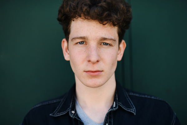 A close-up portrait of young man against green background outdoors, looking at camera.