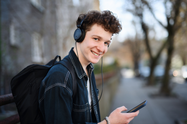 A portrait of young man commuter with headphones standing outdoors in city, using smartphone.