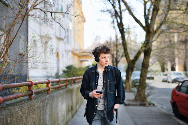 A portrait of young man commuter with headphones walking outdoors in city, using smartphone.