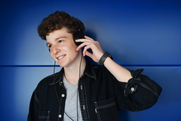 A front view portrait of young man with headphones against blue background outdoors, listening to music.