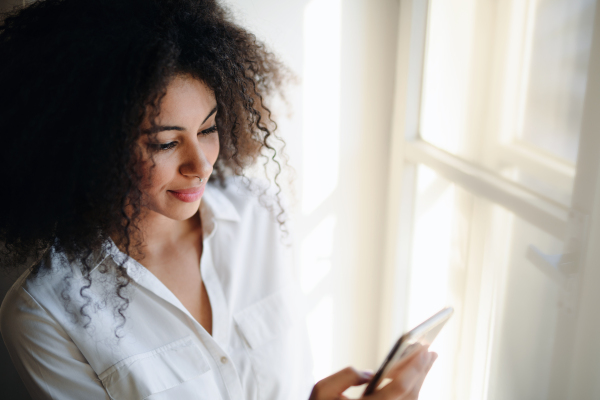 Portrait of happy young woman on window sill indoors at home, using smartphone.