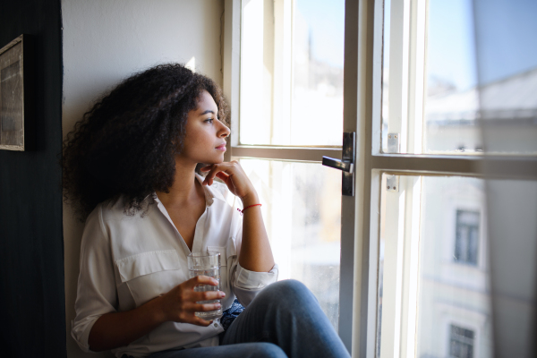 A portrait of young woman sitting on windowsill indoors, looking out and daydreaming.