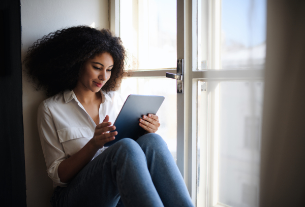 Portrait of young woman sitting on window sill indoors at home, using tablet.