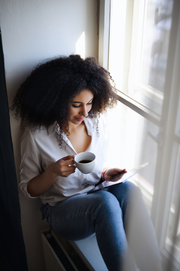 Portrait of happy young woman with coffee indoors at home, using tablet.