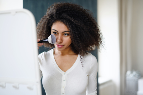 Close-up portrait of young woman with brush indoors at home in front of mirror, applying make-up.