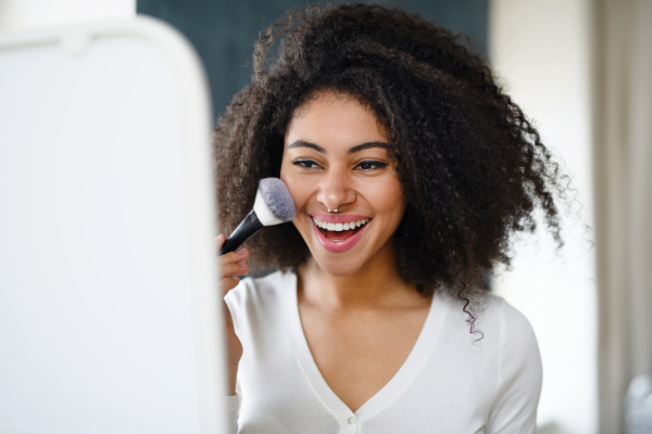 Close-up portrait of young woman with brush indoors at home in front of mirror, applying make-up.