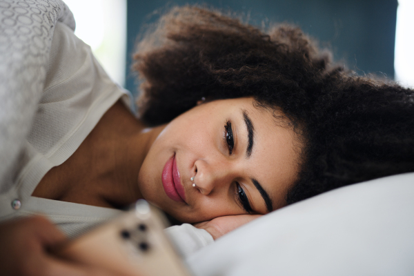 Front view portrait of happy young woman indoors on bed, using smartphone.