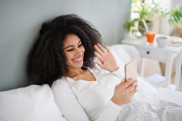 Portrait of cheerful young woman with smartphone indoors on bed, a video call concept.