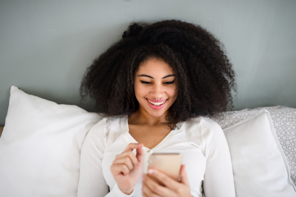 Top view portrait of happy young woman indoors on bed, using smartphone.