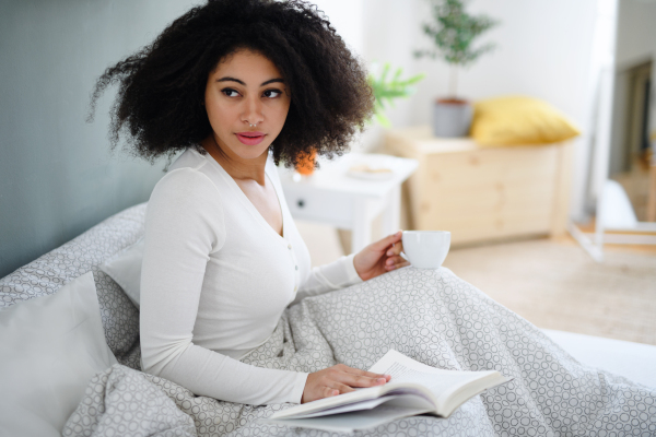 Close-up portrait of happy young woman with book and coffee indoors, relaxing in bed.