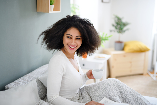 Close-up portrait of happy young woman with book and coffee indoors, relaxing in bed.