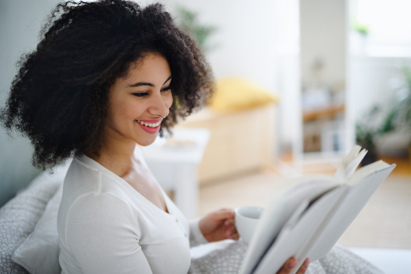 A side view portrait of young woman with book and coffee indoors, relaxing in bed.