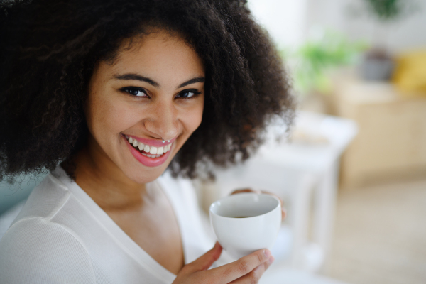 Close-up front view portrait of laughing young woman with coffee indoors, looking at camera.