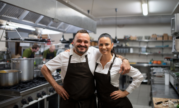 Confident chefs colleagues standing with arms around and looking at a camera in commercial kitchen.