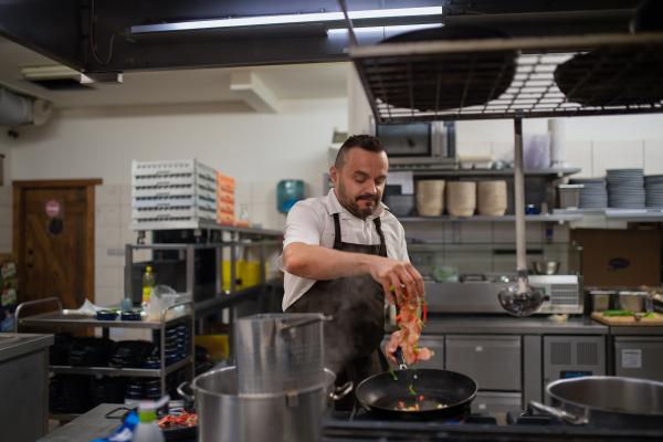 A mature chef adding chopped vehetagles to pan indoors in restaurant kitchen.