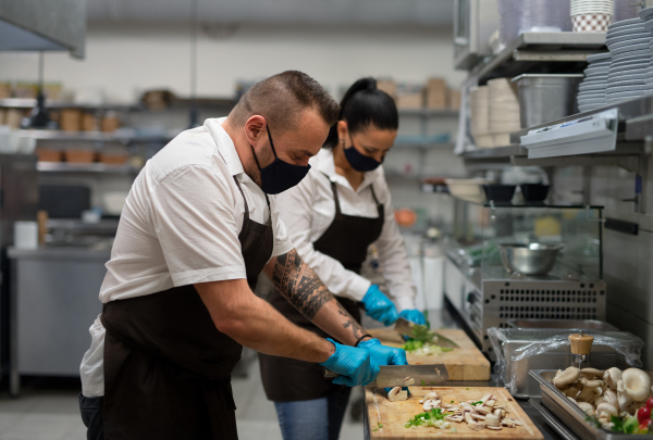 A chef and cook with face masks cutting vegetables indoors in restaurant kitchen, coronavirus concept.