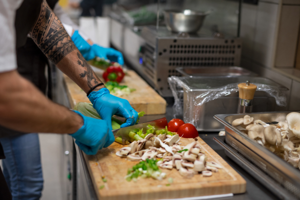 An unrecognizable chef with gloves cutting vegetables indoors in restaurant kitchen.