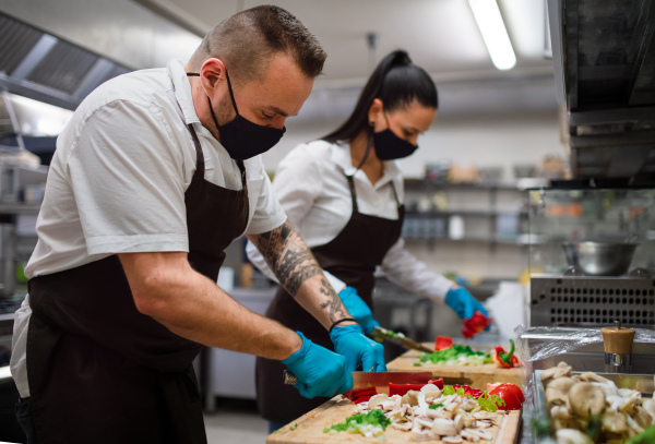 A chef and cook with face masks cutting vegetables indoors in restaurant kitchen, coronavirus concept.