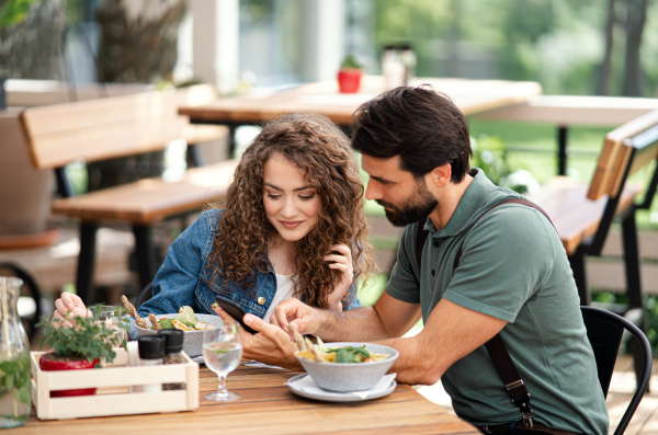 Happy young couple sitting outdoors on terrace restaurant, using smartphone.