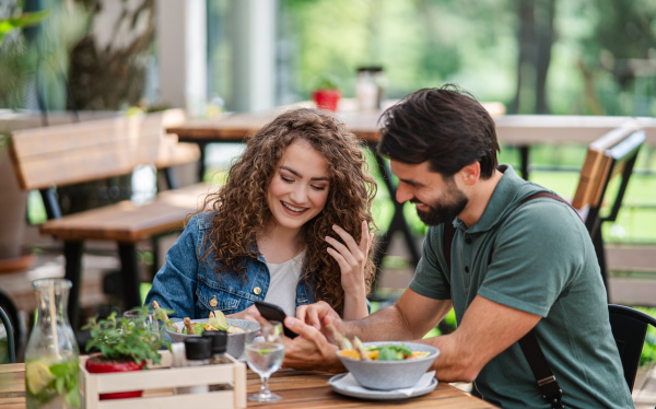 Happy young couple sitting outdoors on terrace restaurant, using smartphone.