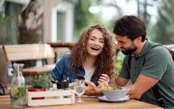 Happy young couple sitting outdoors on terrace restaurant, using smartphone.