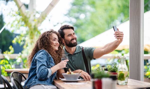 Happy couple sitting outdoors on terrace restaurant, taking selfie with smartphone.