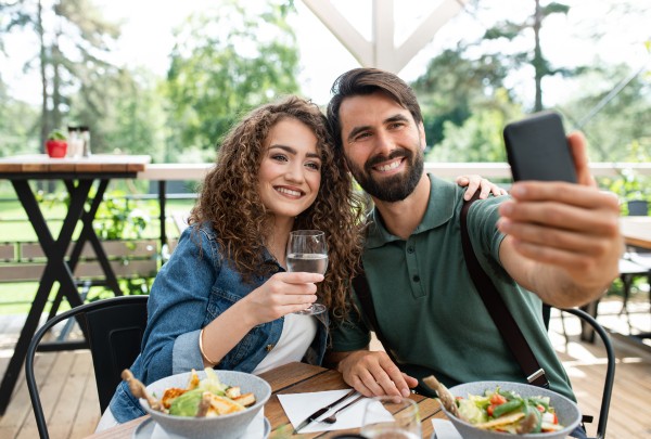 Happy couple sitting outdoors on terrace restaurant, taking selfie with smartphone.