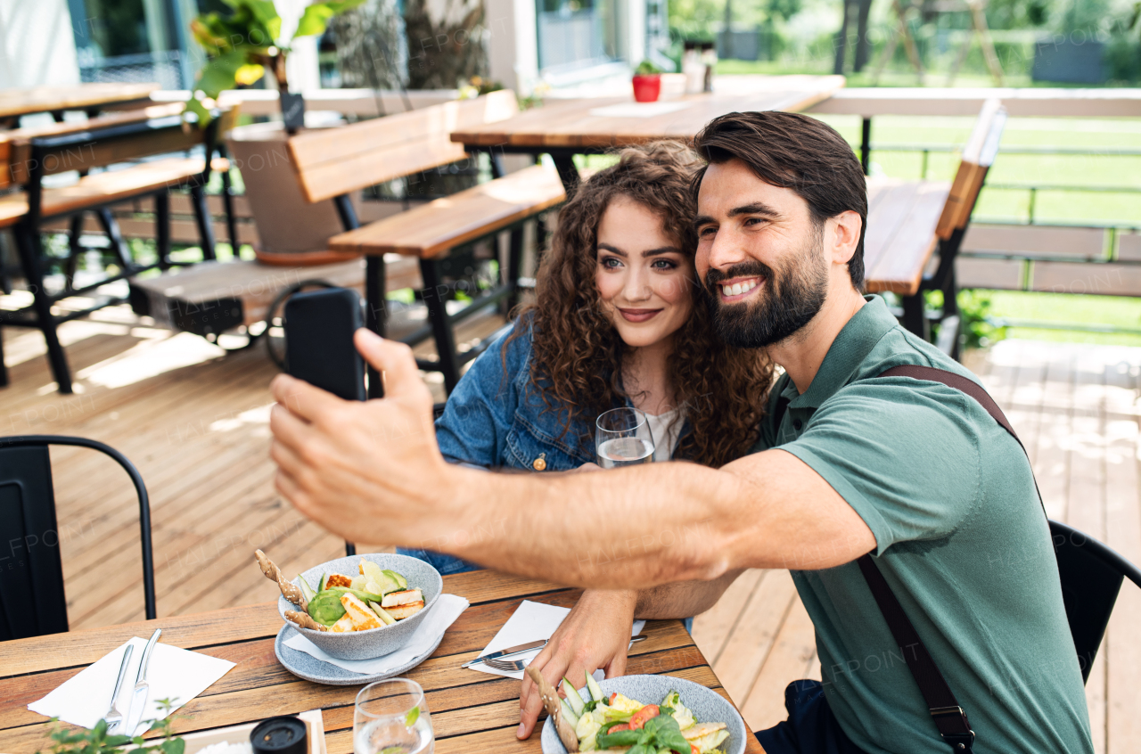 Happy couple sitting outdoors on terrace restaurant, taking selfie with smartphone.