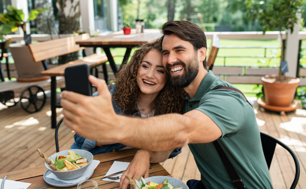 Happy couple sitting outdoors on terrace restaurant, taking selfie with smartphone.