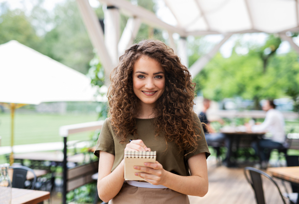 Front view portrait of waitress standing on terrace restaurant, holding order pad.