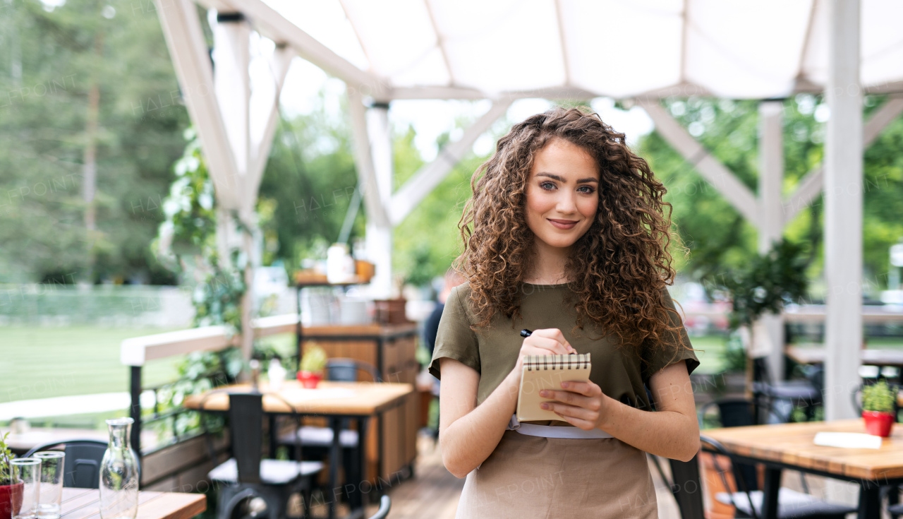 Front view portrait of waitress standing on terrace restaurant, holding order pad.