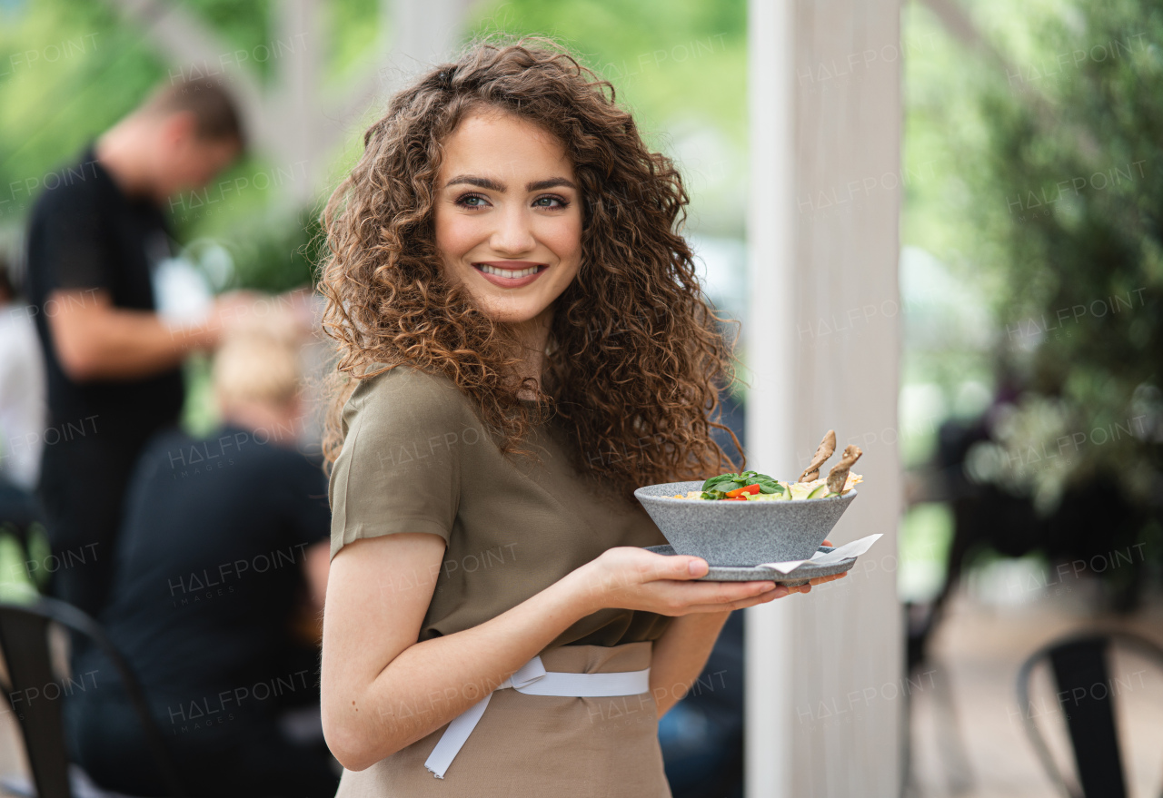 Portrait of waitress with plate standing on terrace restaurant, looking at camera.