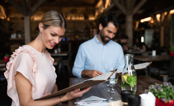 Happy young couple sitting indoors in restaurant, looking at menu.