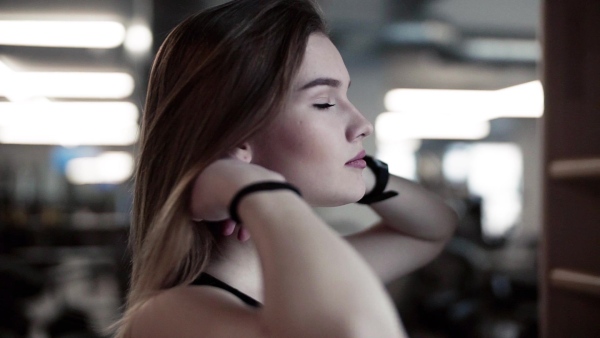 A portrait of a beautiful young girl or woman standing in a gym, doing a ponytail. Slow motion.