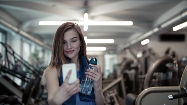 A portrait of young girl or woman with water bottle and smartphone in a gym, taking selfie. Slow motion.