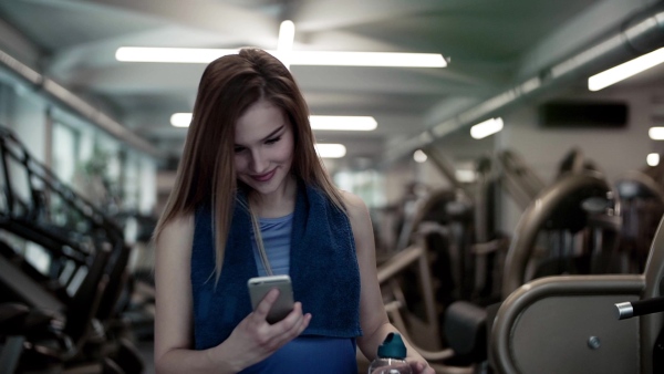 A portrait of young girl or woman with water bottle and smartphone in a gym, taking selfie. Slow motion.