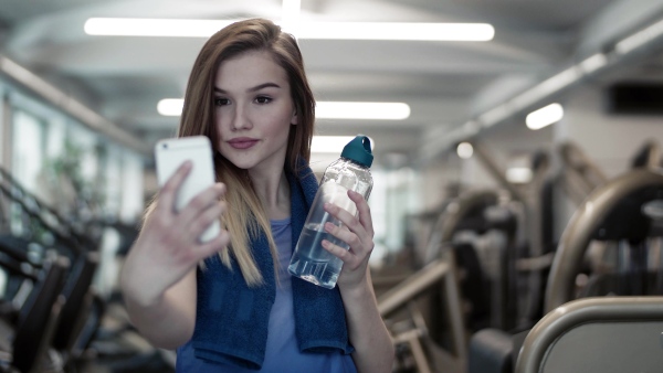 A portrait of young girl or woman with water bottle and smartphone in a gym, taking selfie.