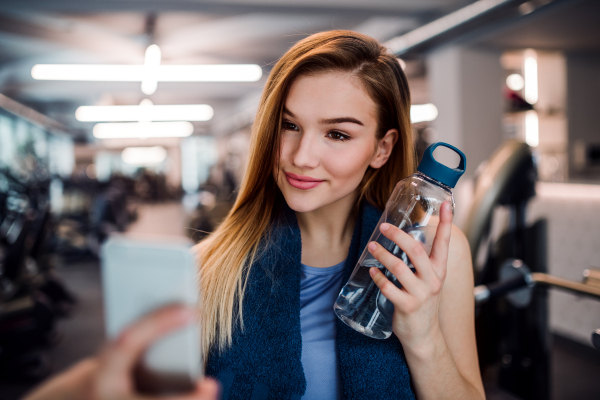 A portrait of young girl or woman with water bottle and smartphone in a gym, taking selfie.